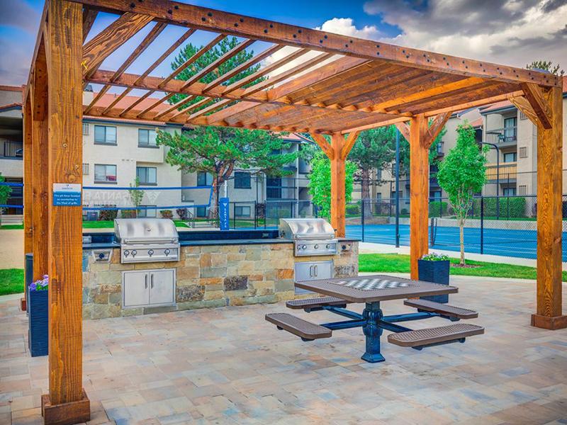 2-BR Apartments in Cottonwood Heights, UT - Santa Fe at Cottonwood - Grill Area with Two BBQs, a Table, and a View of Tennis Courts.